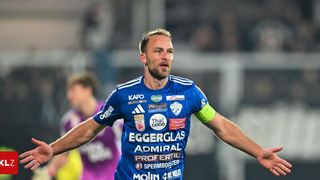 Ein Fußballspieler in einem blauen Trikot feiert. Seine Arme sind weit ausgebreitet. Dahinter beobachtet ein anderer Spieler in einem lila Trikot.