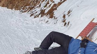 Eine Person liegt auf schneebedecktem Boden, trägt eine blaue Jacke, schwarze Hose und Handschuhe. Hinter ihr befindet sich ein roter Schlitten.