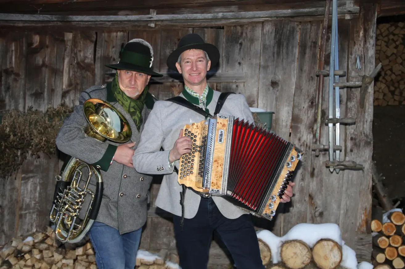 Zwei Männer in traditioneller Kleidung stehen nebeneinander. Ein Mann hält eine Posaune und der andere ein Akkordeon. Sie stehen vor einer Holzwand.