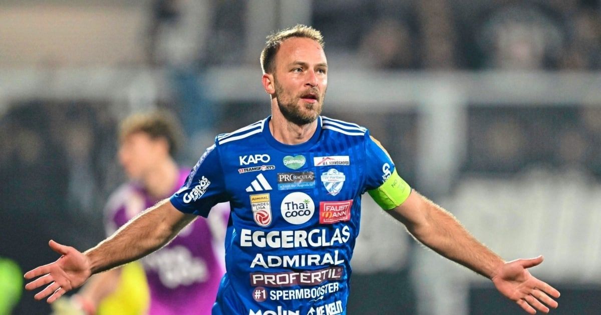 Ein Fußballspieler in einem blauen Trikot feiert auf einem Feld mit ausgestreckten Armen.