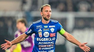 Ein Fußballspieler in einem blauen Trikot feiert auf einem Feld mit ausgestreckten Armen.