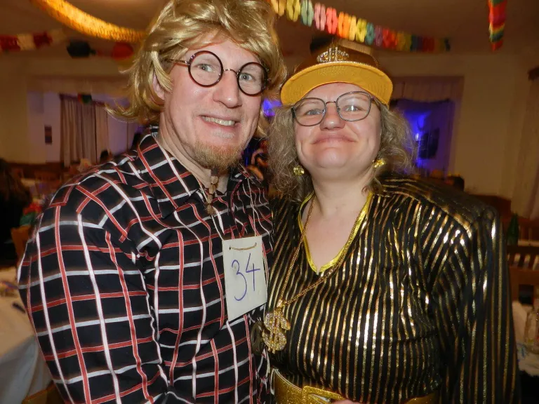 A man and a woman are smiling for a picture at a party. They are wearing costumes. The man is wearing glasses and a checkered shirt, while the woman is wearing a gold dress and a hat. There is a number 34 on a tag on the man's shirt.