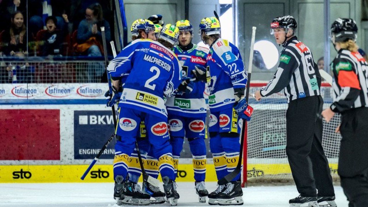 Eine Eishockeymannschaft feiert ein Tor, Spieler tragen blaue Trikots mit verschiedenen Nummern und Sponsoren. Sie stehen auf dem Eis, ein Schiedsrichter ist in der Nähe.