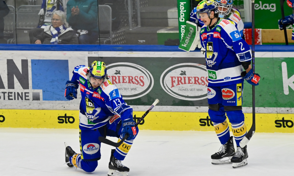 Zwei Eishockeyspieler auf dem Eis, einer kniet mit einem Eishockeyschläger, der andere steht mit einem Eishockeyschläger, beide tragen blaue Uniformen mit Nummern und Helmen. Dahinter eine Glasbarriere mit Werbetafeln und sitzenden Personen.