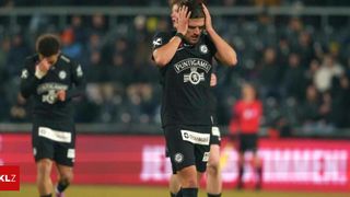 Ein Fußballspieler in einem schwarzen Trikot mit der Aufschrift Puntigames hält sich den Kopf. Hinter ihm laufen andere Spieler. Im Hintergrund sind verschwommene Menschen auf den Tribünen zu sehen.