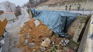 Ein Baustellenbereich mit einem grünen Planen bedeckt. Steine, Erde und Schutt sind verstreut. Zwei Feuerwehrleute stehen in der Nähe.