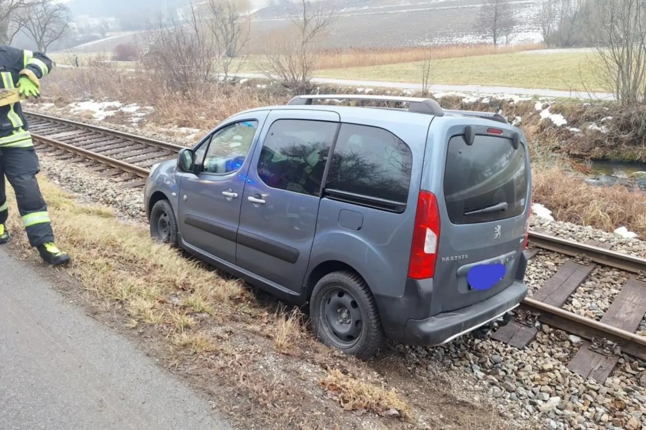 Ein grauer Van ist am Rande einer Bahnlinie geparkt. Er hat ein Dachgepäckträger und ein Nummernschild, das 'PARTNER' liest. Die Rückseite des Vans ist beschädigt.