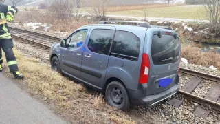 Ein grauer Van ist am Rande einer Bahnlinie geparkt. Er hat ein Dachgepäckträger und ein Nummernschild, das 'PARTNER' liest. Die Rückseite des Vans ist beschädigt.