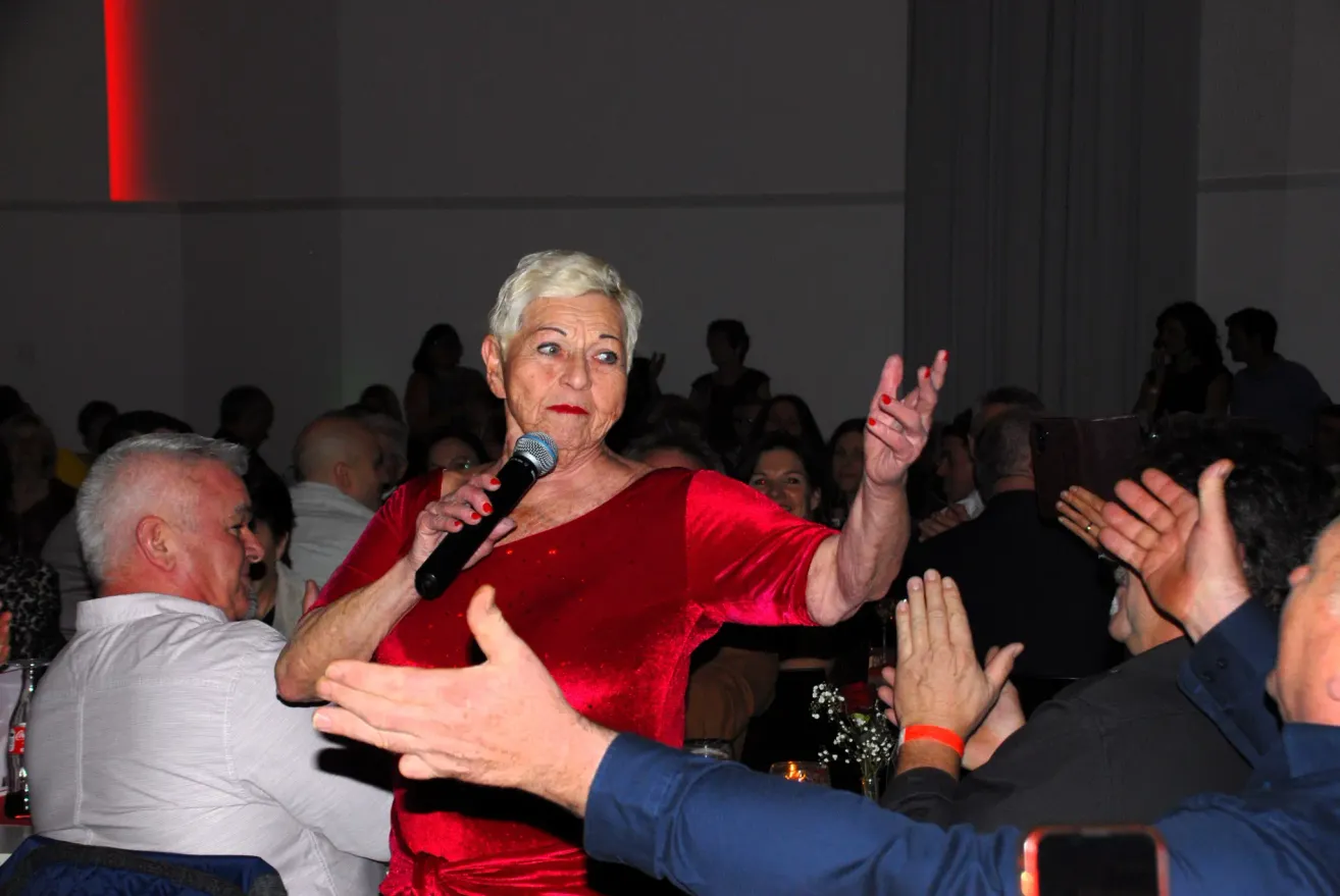 Eine ältere Frau in einem roten Kleid hält ein Mikrofon und singt möglicherweise. Sie ist von klatschenden Menschen umgeben. Einige Personen sind verschwommen. Auf dem Tisch steht eine Vase mit Blumen.