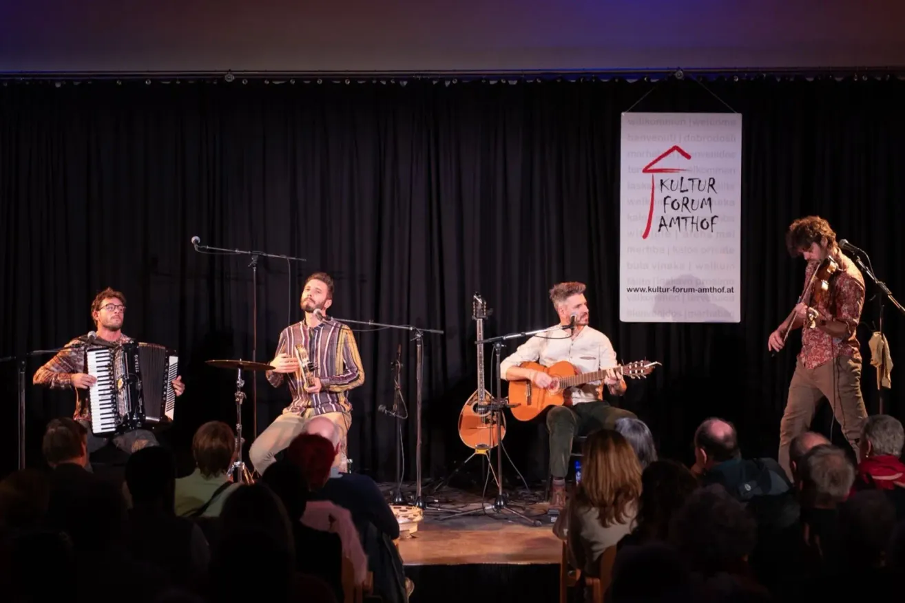 Eine kleine Band tritt auf der Bühne im Kultur Forum Amhof auf, mit einem Publikum vor ihnen. Ein Mann spielt Akkordeon, ein anderer Gitarre und ein Dritter Schlagzeug.