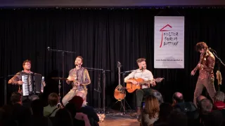 Eine kleine Band tritt auf der Bühne im Kultur Forum Amhof auf, mit einem Publikum vor ihnen. Ein Mann spielt Akkordeon, ein anderer Gitarre und ein Dritter Schlagzeug.