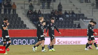 Drei Fußballspieler gehen auf einem Feld. Zwei von ihnen halten sich an den Händen. Hinter ihnen ist ein Stadion mit Zuschauern zu sehen. Ein Werbebanner ist auf dem Feld vorhanden.