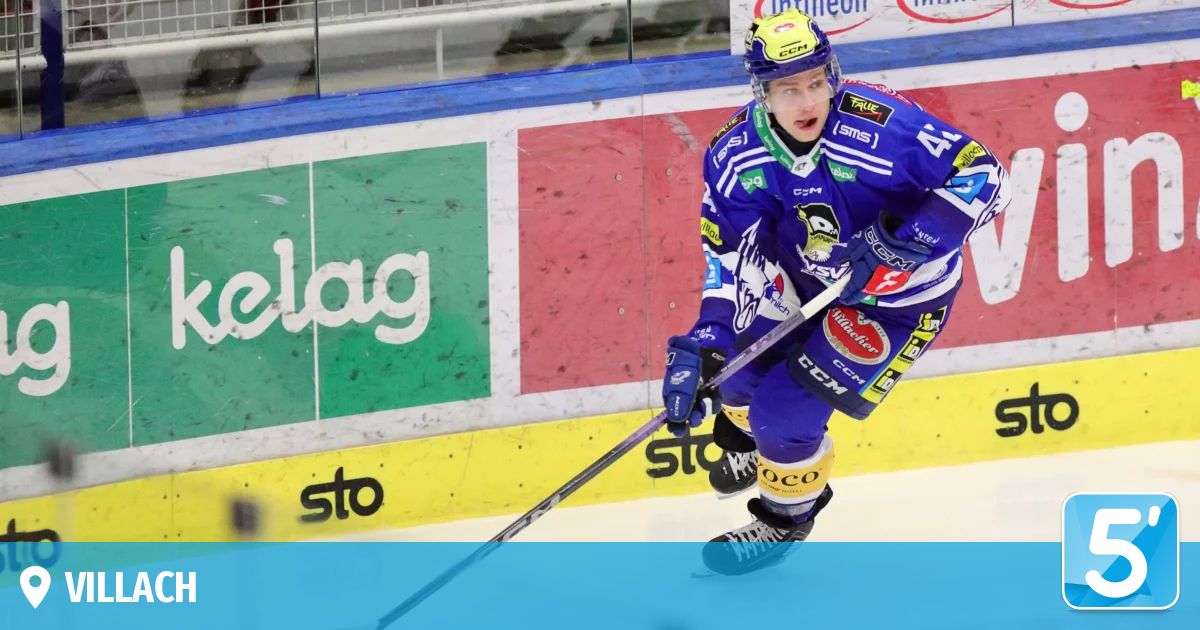 Ein Eishockeyspieler in einem blauen Trikot ist auf dem Eis, hält einen Schläger und schaut nach links. Hinter ihm ist eine Glasbarriere mit Werbung.