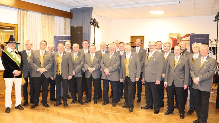 A group of formally dressed men, likely a choir, stand in a room with wooden floors. They wear matching suits with yellow vests and ties. Behind them, a banner and speakers are visible.