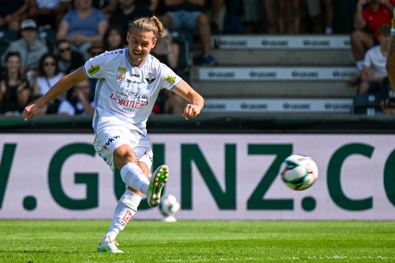 Ein Fußballspieler in einem weißen Trikot tritt einen Ball auf einem Feld mit einem Banner, das 'GUNZ' steht. Der Spieler ist in Aktion, und es gibt Zuschauer im Hintergrund.