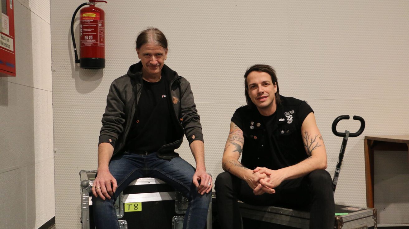 Zwei Männer sitzen auf einer Aufbewahrungsbox mit einem Feuerlöscher im Hintergrund. Beide tragen schwarze T-Shirts, Jeans und haben Tattoos.