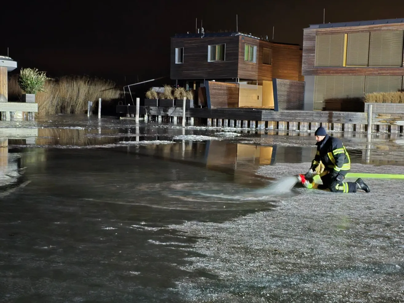 A firefighter is kneeling on the ice, spraying water from a hose. There are houses and a pier in the background.