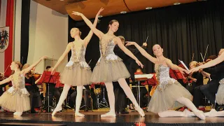 Three ballerinas perform on stage, one kneeling, one standing, and one leaping, with musicians and music stands in the background.
