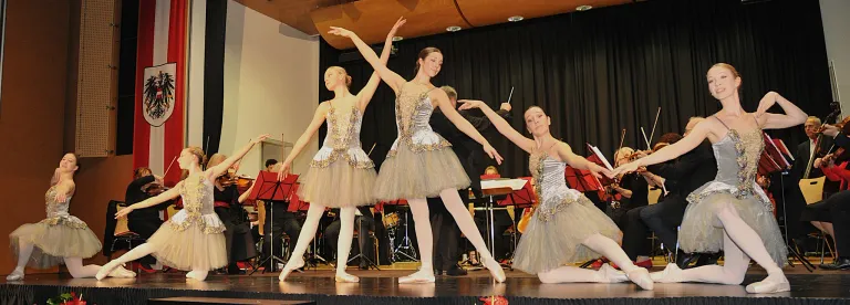 Drei Ballerinen treten auf der Bühne auf, eine kniet, eine steht und eine springt, mit Musikern und Notenständern im Hintergrund.