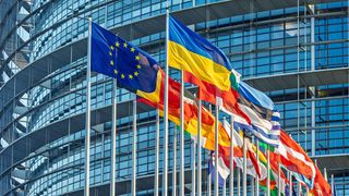 Flags of various nations wave in the wind outside a modern, glass-walled building, likely a governmental institution.