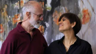 An elderly man wearing glasses and a maroon polo stands beside a smiling woman in a black blouse, both looking upward against a vibrant abstract painting.