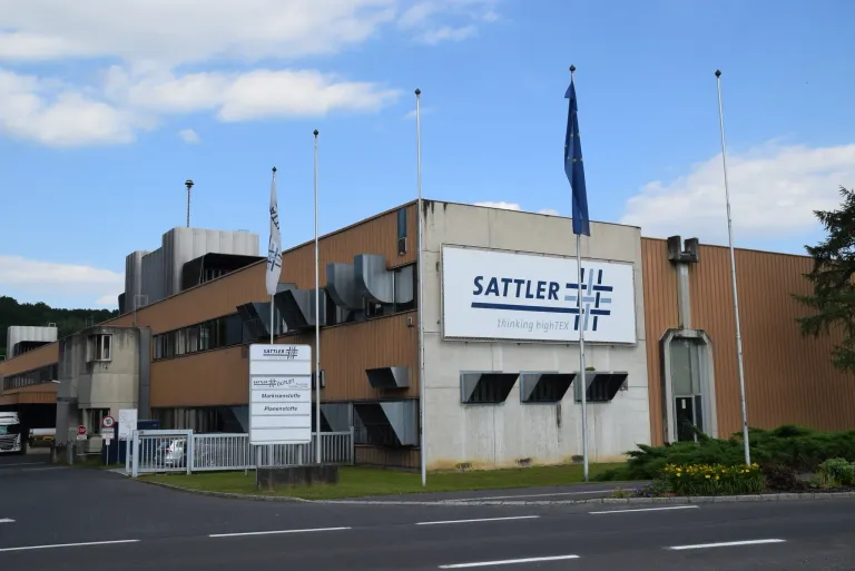 Ein SATTLER-Industriegebäude hat ein weißes Schild mit dem Firmenlogo und dem Text 'thinking highTEX'. Zwei Fahnen stehen auf beiden Seiten des Eingangs, und ein weißes Schild steht auf dem Rasen vor dem Gebäude.