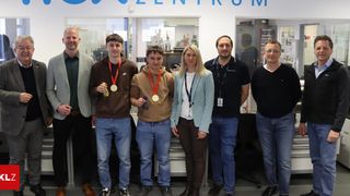 A group of people stand in a room with a white wall and the word Zentrum on it. Two men wear medals around their necks.