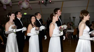A group of people are standing on a wooden floor. They are wearing formal attire, holding flowers, and smiling. They are probably participating in a wedding ceremony. In the background, there is a table with chairs, and some people are sitting. The ceiling has a few lamps, and a few balloons are hanging.