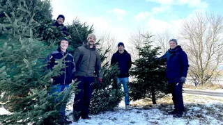 A group of people stand in a snowy area, holding Christmas trees. They wear winter clothes and seem to be posing for a photo. Behind them, there are trees with no leaves.