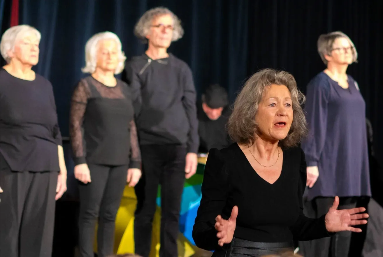 A group of people in black clothing on stage, one speaking with hands gestures. Behind them, a blurred image of a colorful backdrop.