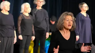 A group of people in black clothing on stage, one speaking with hands gestures. Behind them, a blurred image of a colorful backdrop.