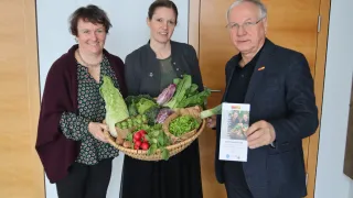 Three individuals stand close to each other, smiling. A woman on the left holds a basket filled with various vegetables. Another woman in the middle holds a basket with green vegetables, and a man on the right holds a card.
