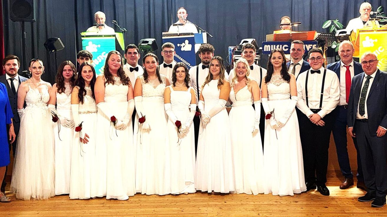 A group of people in white dresses and white gloves are standing on stage with musical instruments in the background.