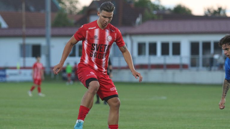 A soccer player in a red uniform runs on a field, looking down. The jersey says 'SVETA GROUP' and has sponsors' logos. In the background, a building with a red roof is visible.