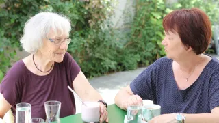 Two women are sitting at a table, smiling, with drinks in front of them. The woman on the left has white hair and is wearing glasses. The woman on the right is wearing a watch.