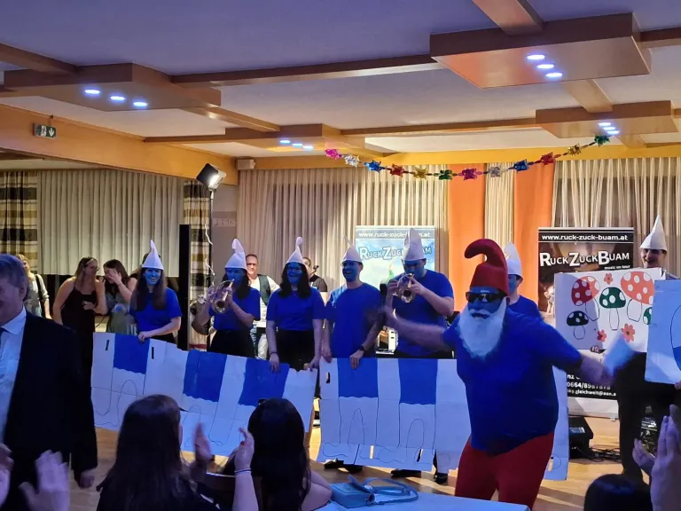 Eine Gruppe von Menschen mit blauer Gesichtsbemalung und Kostümen führt eine synchronisierte Darbietung in einem dekorierten Raum auf, mit einer Person in einem Weihnachtsmann-Kostüm im Vordergrund.