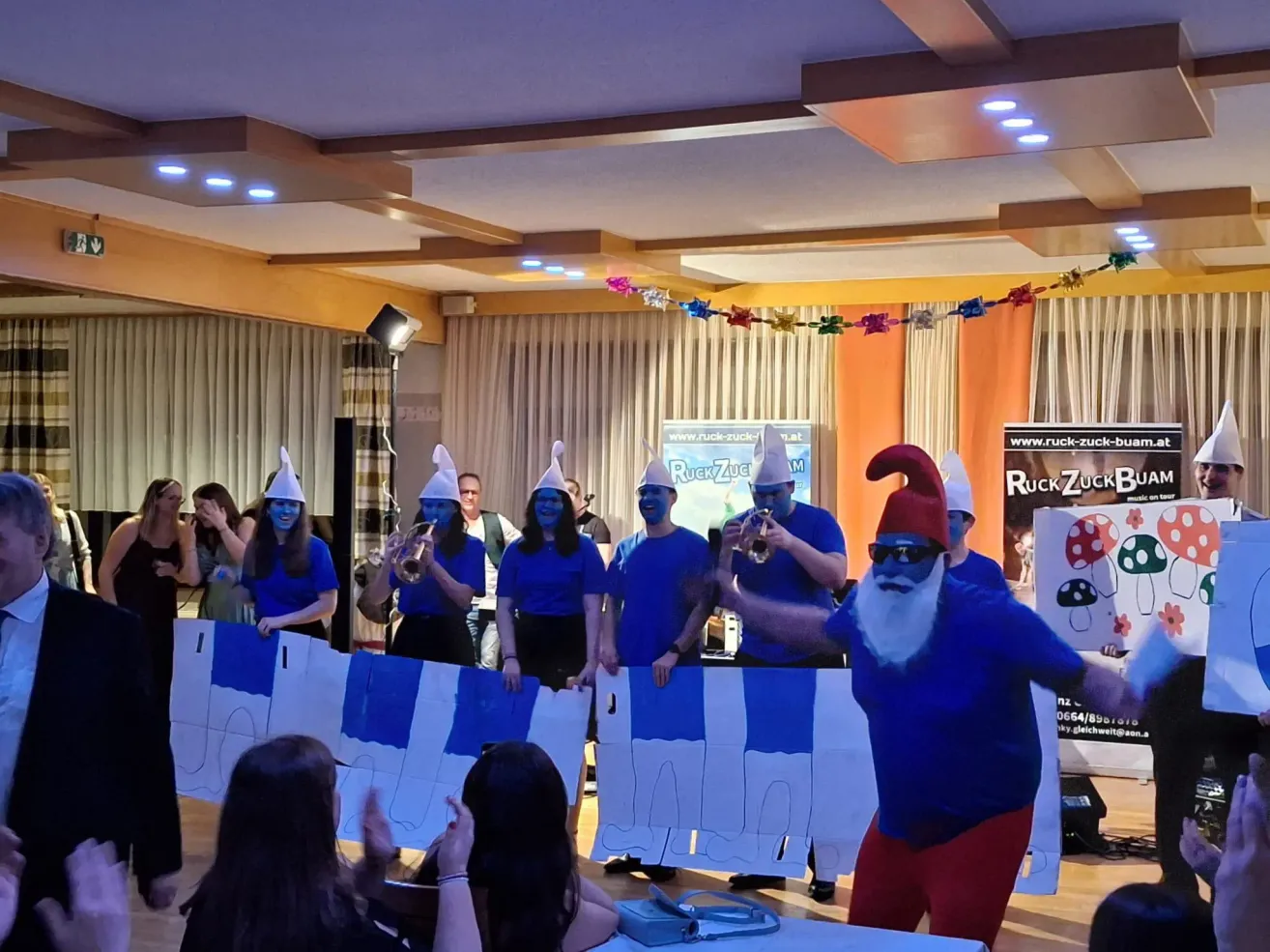 Eine Gruppe von Menschen mit blauer Gesichtsbemalung und Kostümen führt eine synchronisierte Darbietung in einem dekorierten Raum auf, mit einer Person in einem Weihnachtsmann-Kostüm im Vordergrund.