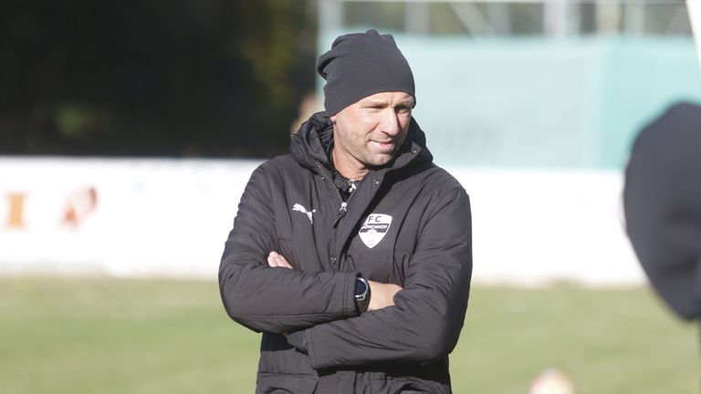 A man wearing a black jacket and a beanie stands on a field with his arms crossed.