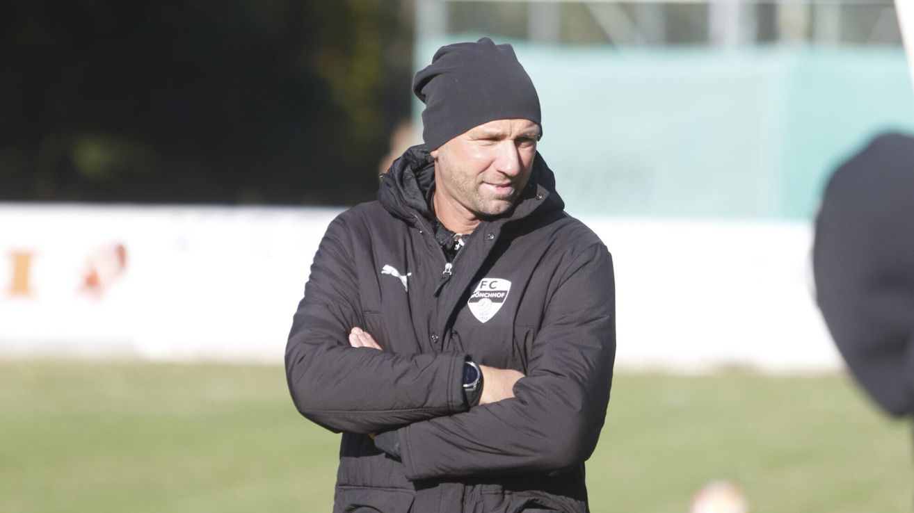 Ein Mann in einer schwarzen Jacke und einer Mütze steht auf einem Feld mit verschränkten Armen.