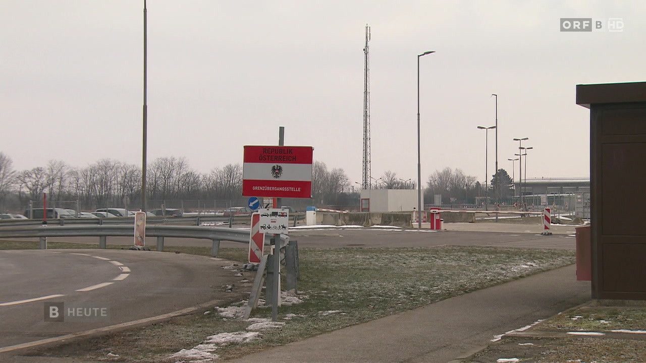 Ein Grenzübergangskontrollpunkt mit einem roten und weißen Schild, auf dem 'Republik Osterreich Grenzübergangsstelle' steht. Es sind hohe Straßenlaternen, Bäume und ein Gebäude sichtbar.
