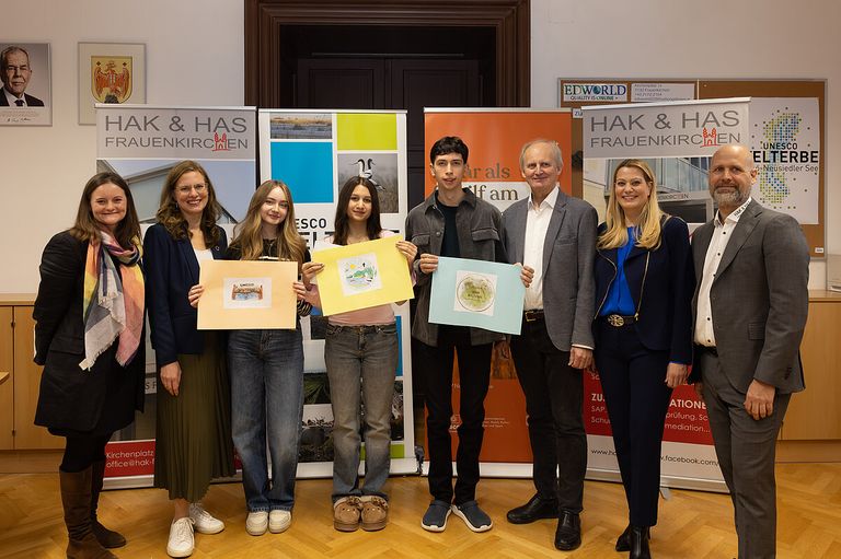 Fünf Personen stehen in einem Raum und posieren für ein Foto vor Bannern im Hintergrund. Sie halten bunte Papiere hoch. Zwei Frauen links tragen Brillen, die mittlere Figur ist ein Junge. Rechts stehen zwei Männer in Anzügen.