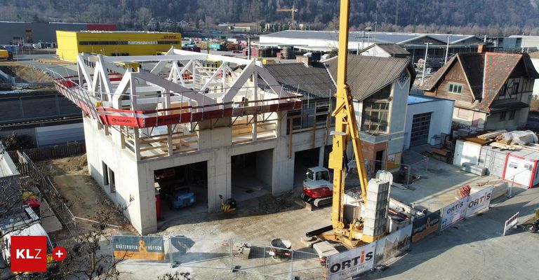 Luftaufnahme einer Baustelle mit einem gelben Kran, einem teilweise fertiggestellten Gebäude und verschiedenen Baufahrzeugen und Materialien an einem klaren Tag.
