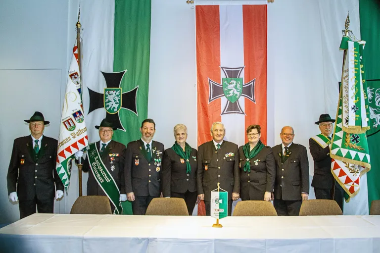 Six people in uniform stand in front of a table with flags in the background. Two flags have a cross with a shield in the center.