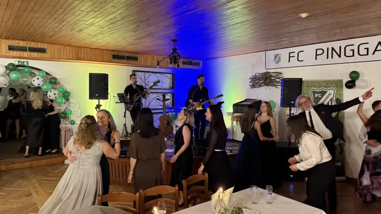 Zwei Männer spielen Gitarren auf der Bühne, während zwei Frauen vor ihnen tanzen. Auf beiden Seiten der Bühne befinden sich zwei Lautsprecher. Ein Banner mit dem Text FC ist an der Wand hinter der Bühne.