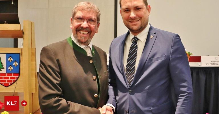Zwei Männer in formeller Kleidung lächeln und schütteln Hände. Der Mann auf der linken Seite trägt eine traditionelle grüne Weste über einem weißen Hemd. Der Mann auf der rechten Seite ist in einem blauen Anzug und Krawatte.