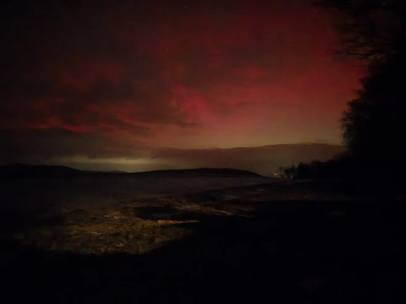 Der Nachthimmel wird von leuchtenden roten und orangefarbenen Tönen beleuchtet, wahrscheinlich verursacht durch das Nordlicht. Ein ruhiger Wasserkörper spiegelt die Farben des Himmels wider. Bäume und Hügel sind in der Ferne kaum sichtbar.