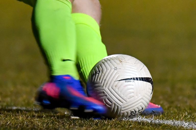 Ein Fußballspieler in neongrünen Socken und blauen Stollenschuhen tritt einen weißen Fußball mit Nike-Logo auf einem Rasenfeld.