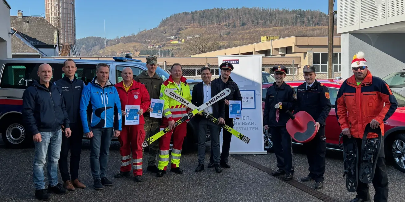 Eine Gruppe von Männern, darunter Polizisten und Rettungskräfte, steht vor einem Gebäude mit einem Banner. Sie halten Urkunden und Skistöcke.