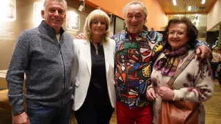 Four people stand close together for a photo. The man on the left wears a gray sweater. The central figure is a woman in a white blazer. The man next to her wears a colorful, patterned shirt. The woman on the right wears glasses and a scarf. They are all smiling.