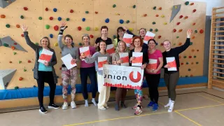A group of women poses for a photo in front of a climbing wall with colorful grips. They hold certificates and a banner with the word Sport Union.